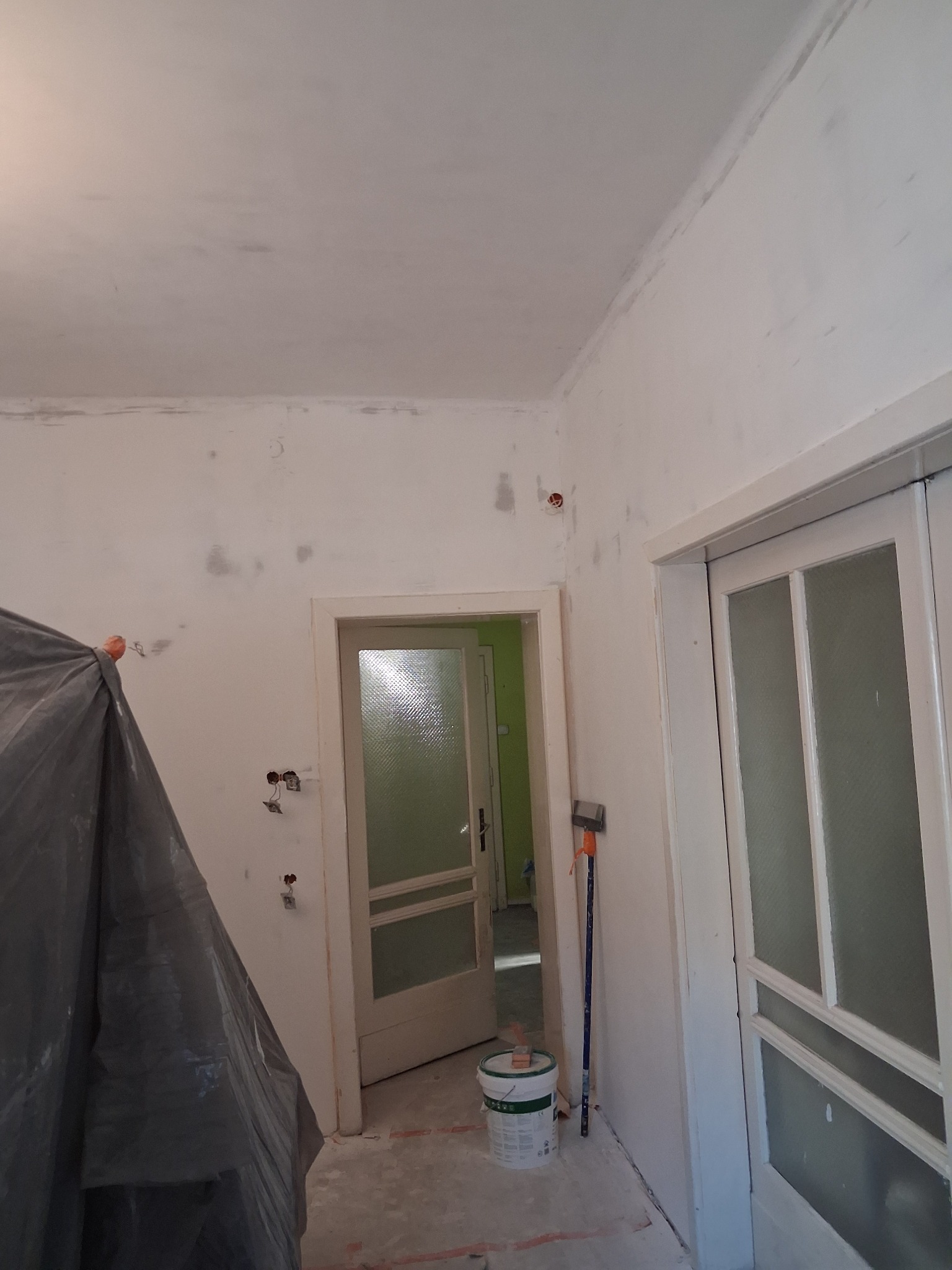 Pokój w trakcie szpachlowania: ściany z widocznymi łatami szpachli, zakryte meble, otwarte drzwi do kolejnego pomieszczenia, wiadro z farbą i wałek malarski.