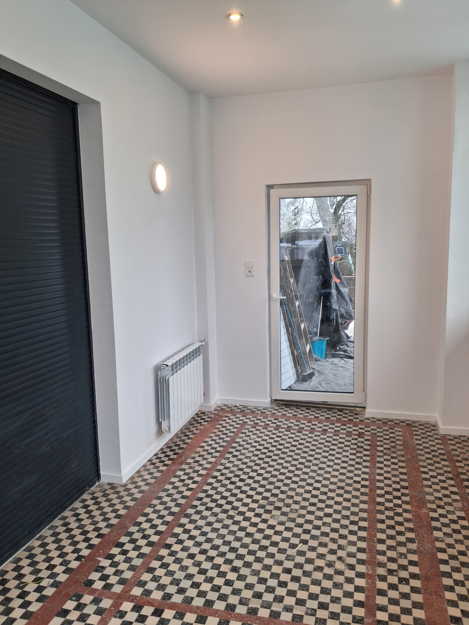 Białe ściany po malowaniu, czarne drzwi z żaluzjami, wzorzysta podłoga z płytek i grzejnik w odnowionym wnętrzu. Widok na zewnątrz przez przeszklone drzwi.