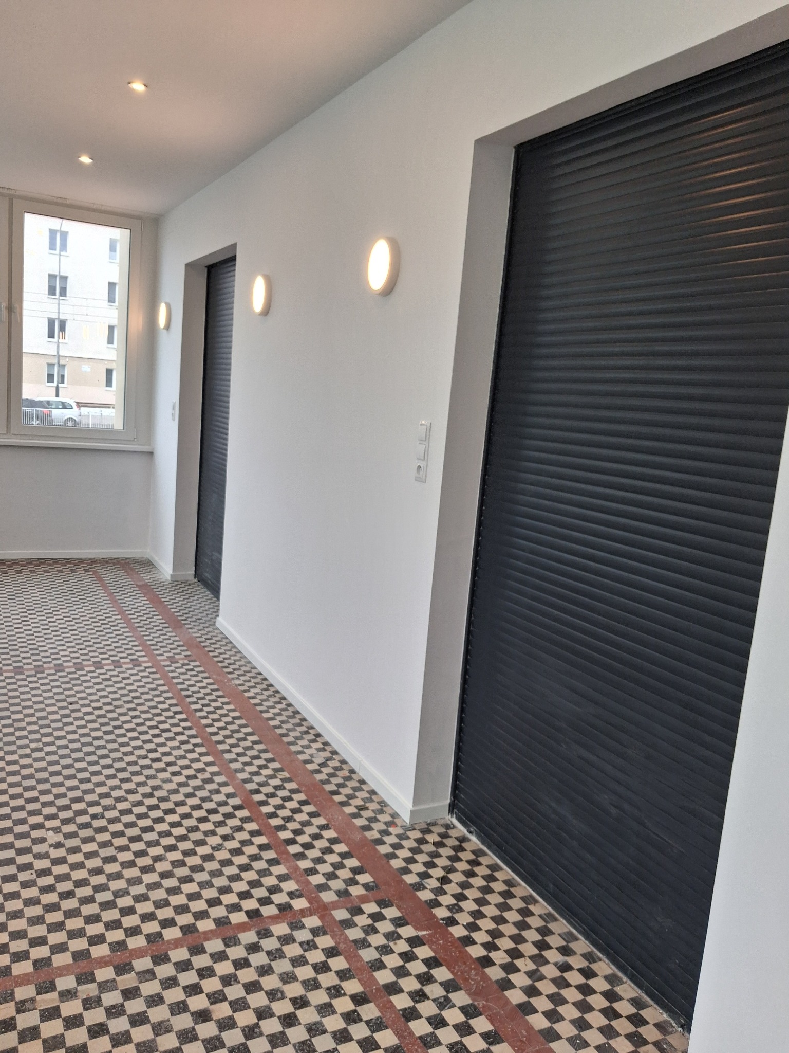 Białe ściany, czarne rolety i wzorzysta podłoga w odnowionym korytarzu. Widoczne detale wykończenia i oświetlenie. Charakterystyczna szachownica na posadzce.