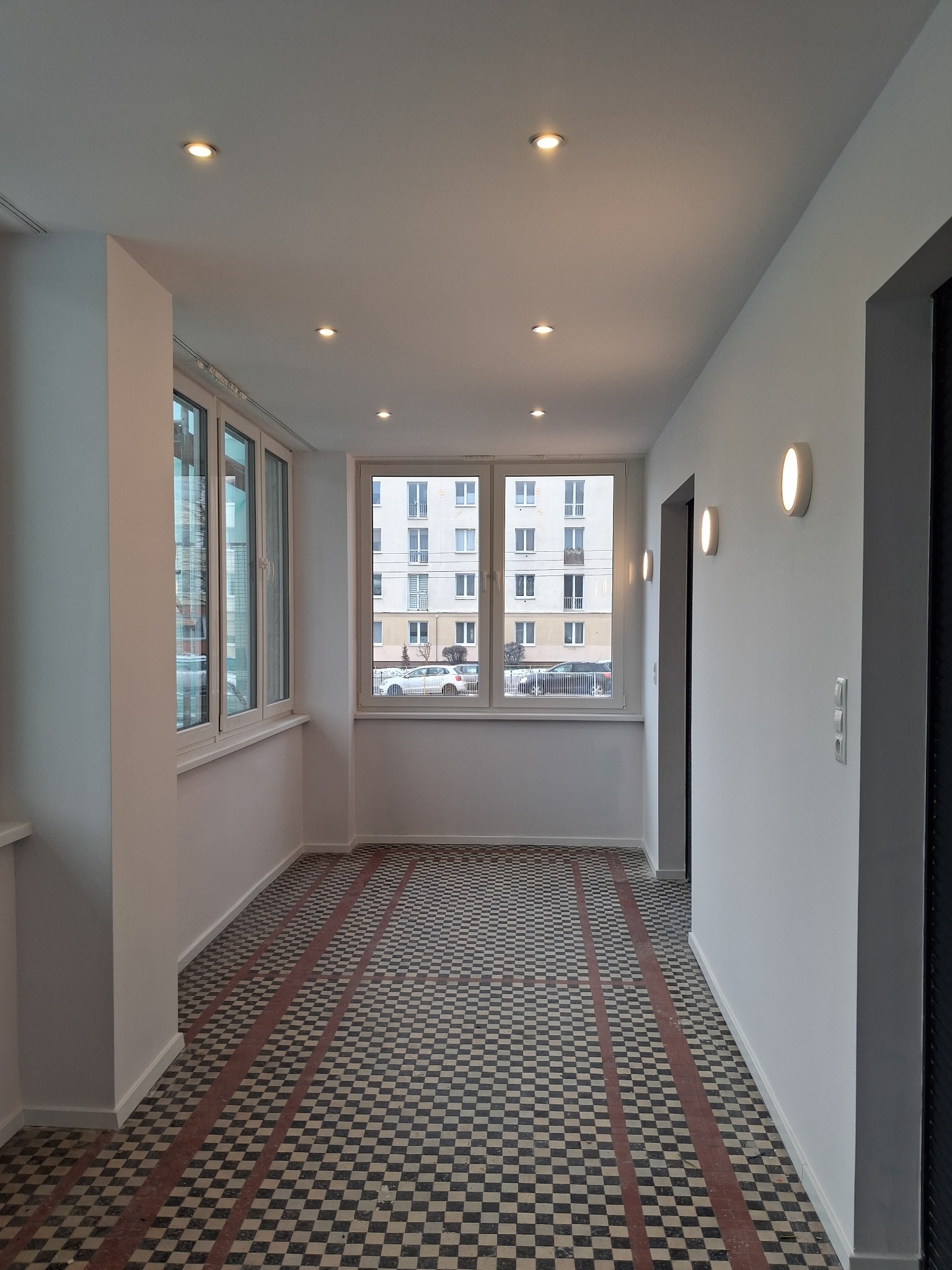 Korytarz po remoncie z białą ścianą, oświetleniem punktowym i podłogą w szachownicę z czerwonymi pasami. Widok na okno z budynkiem w tle.