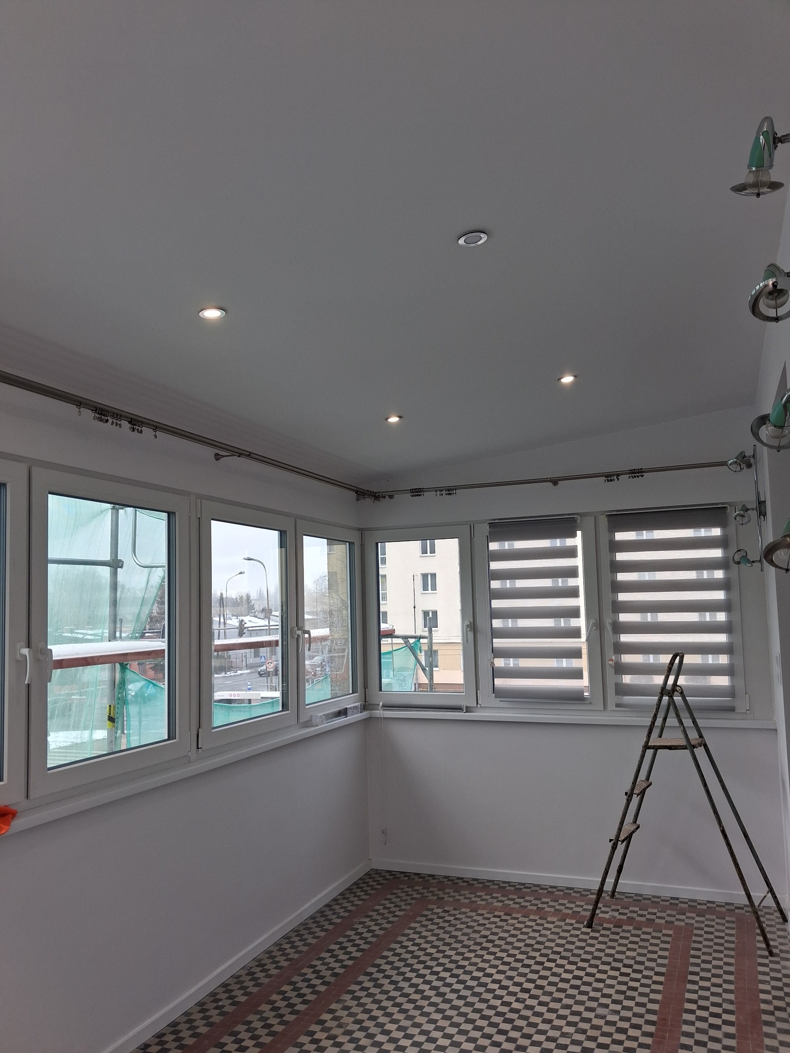 Białe, odnowione wnętrze balkonu z oknami, roletami i wzorzystą podłogą. Widoczne detale wykończeniowe i drabina malarska.