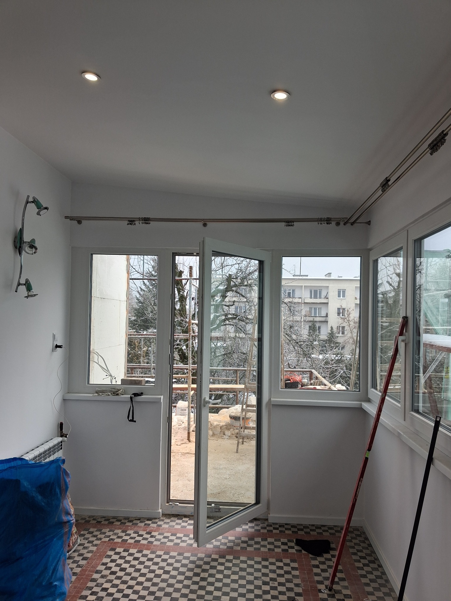 Jasne wnętrze balkonu po remoncie: białe ściany, okna z widokiem na zimowy krajobraz, wzorzysta podłoga i elementy dekoracyjne. Widoczne narzędzia i materiały budowlane.