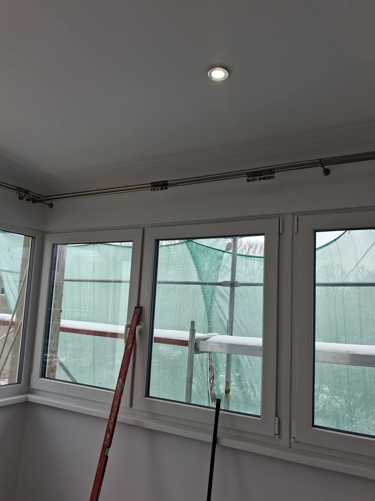 Białe okna z metalowymi karniszami w jasnym pokoju. Widoczne rusztowania za oknem, sugerujące prace wykończeniowe. Ustawiony poziomica przy oknie.