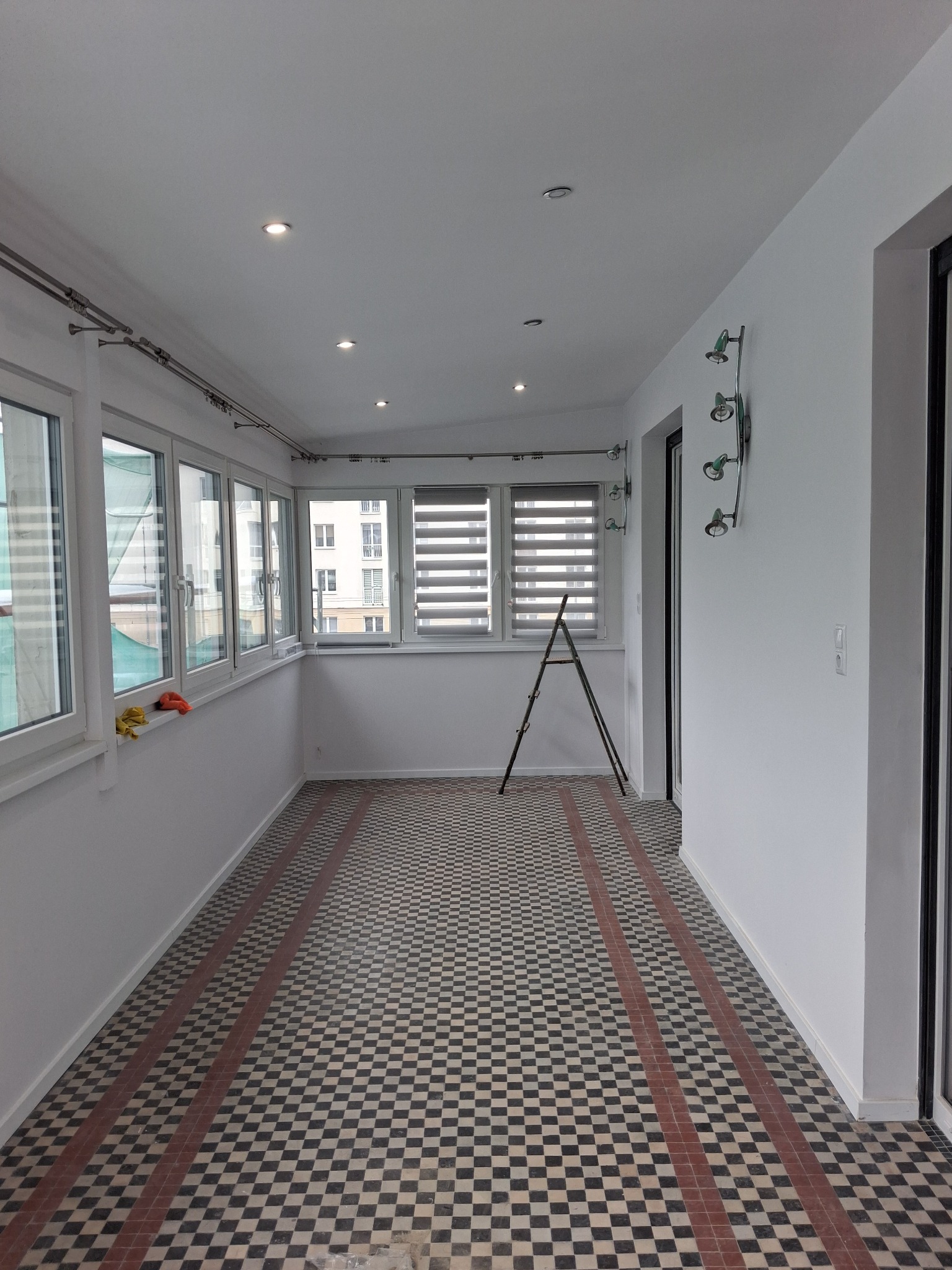 Wnętrze przeszklonego balkonu z czarno-białą podłogą w szachownicę. Widoczne okna, drabina oparta o ścianę, oświetlenie sufitowe i dekoracyjna lampa ścienna.