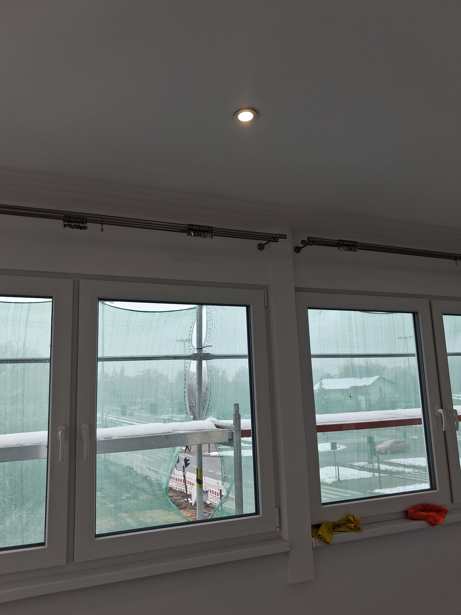 Białe okna z metalowymi karniszami w nowym wnętrzu, zabezpieczone siatką budowlaną. Widok na ulicę z rusztowaniami. Ścierki na parapecie.