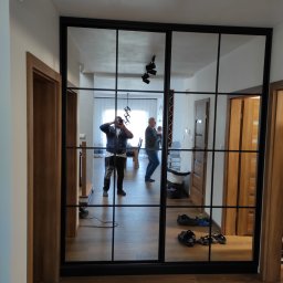 NKmeble - Nowoczesna szafa wnękowa z czarnymi profilami i lustrzanymi drzwiami przesuwnymi, odbijająca wnętrze mieszkania i osoby.