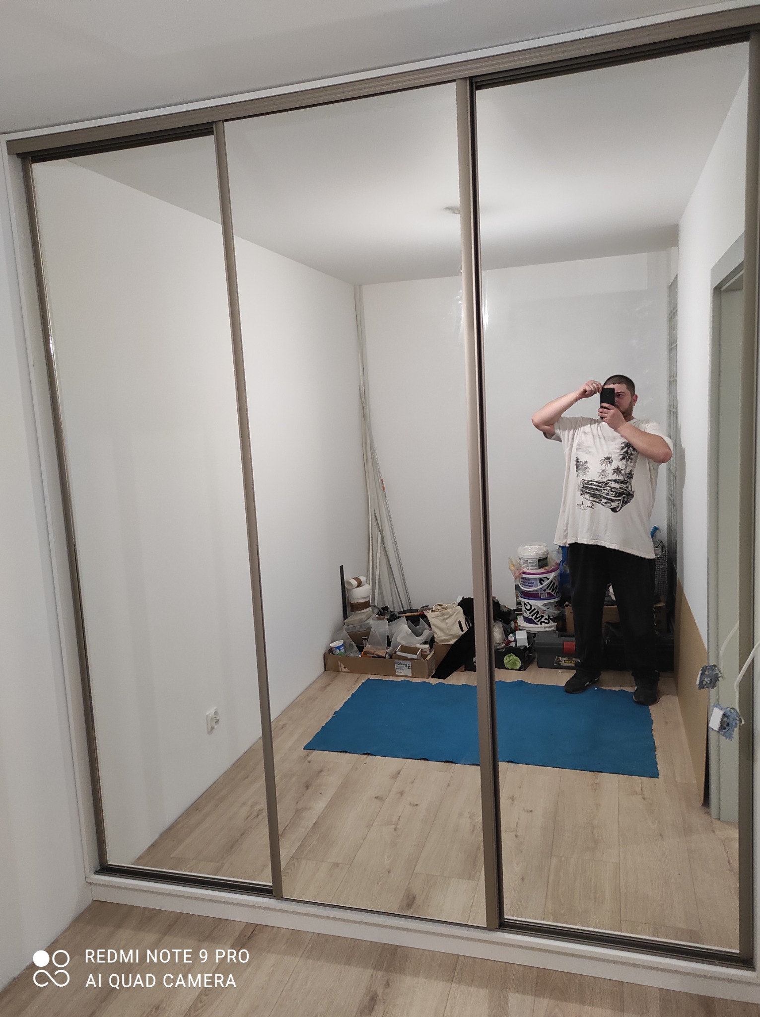 Trzydrzwiowa szafa wnękowa z lustrami, odbijająca wnętrze pokoju z nieporządkiem i osobą robiącą zdjęcie. Widoczne detale wykończenia i podłoga.