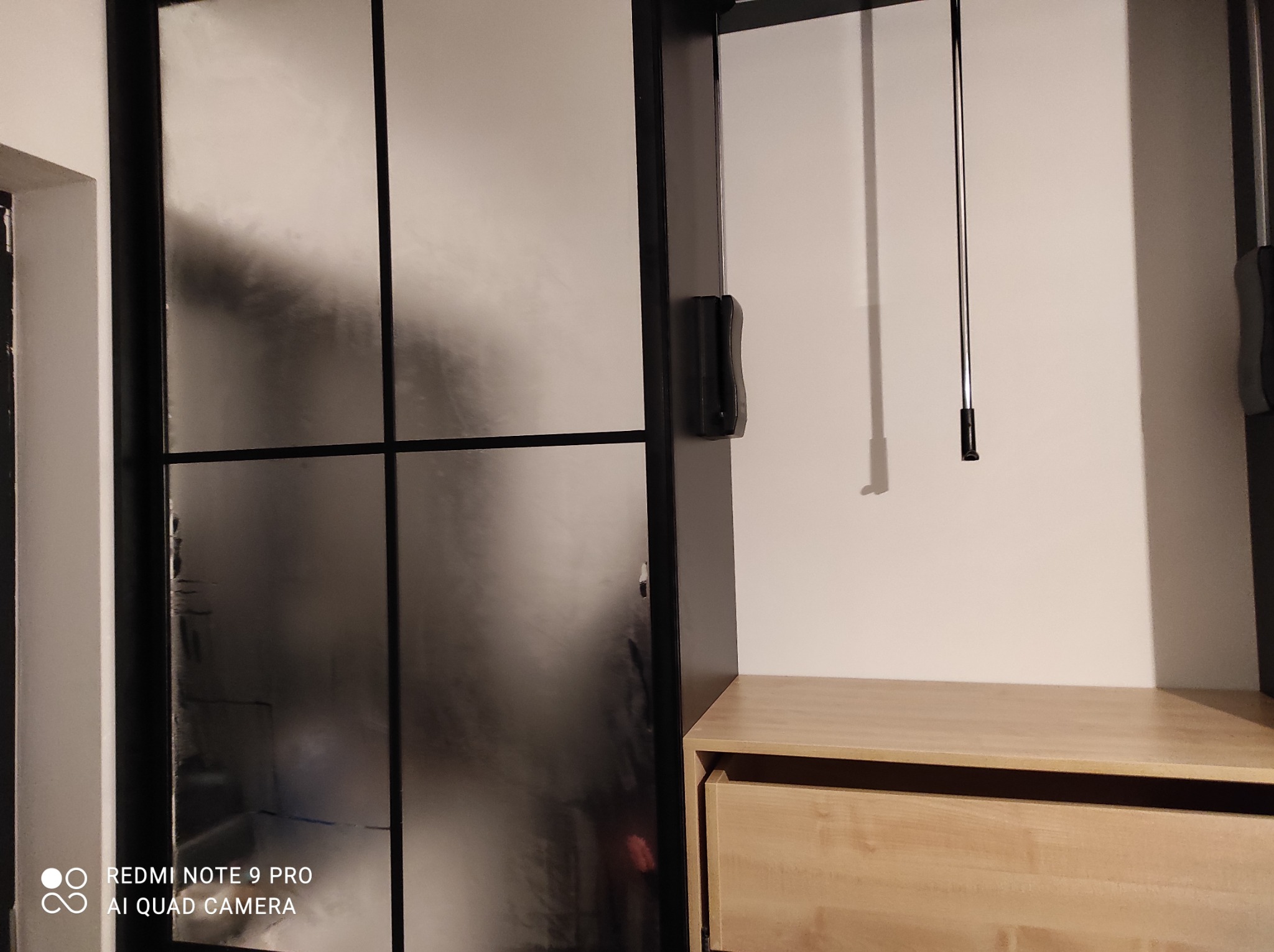 Nowoczesna szafa wnękowa z przesuwnymi drzwiami w stylu loft, z czarnymi szprosami i matowym szkłem, uzupełniona drewnianą komodą z szufladami.