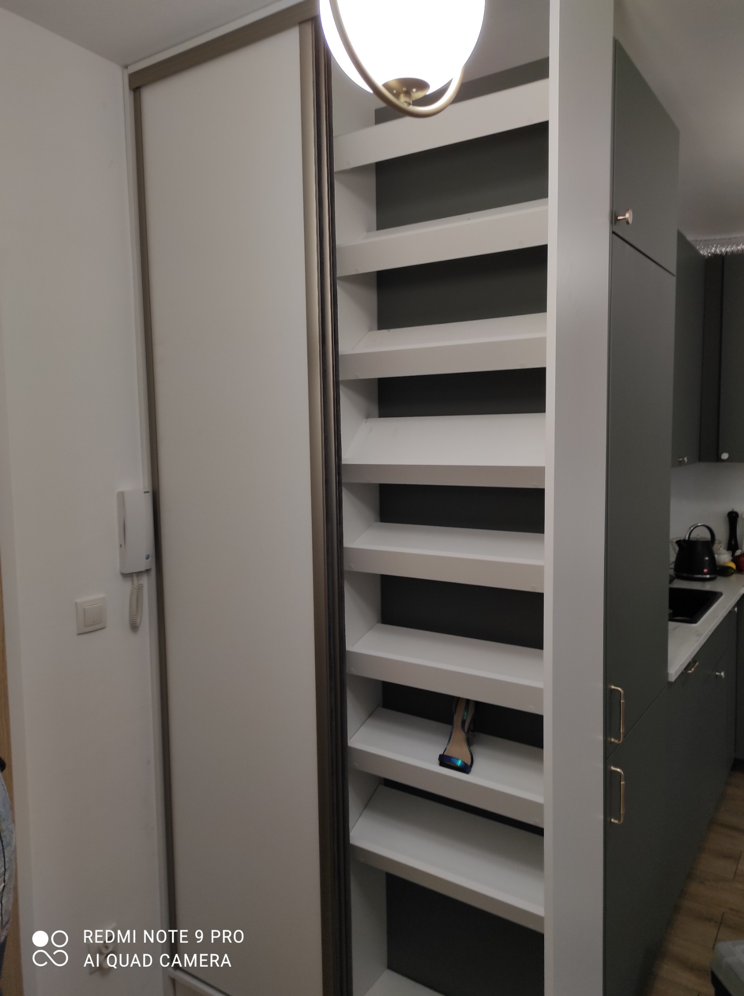 Biała szafa wnękowa z przesuwnymi drzwiami i otwartymi półkami, zintegrowana z zabudową kuchenną w odcieniach szarości. Na jednej z półek leży but.