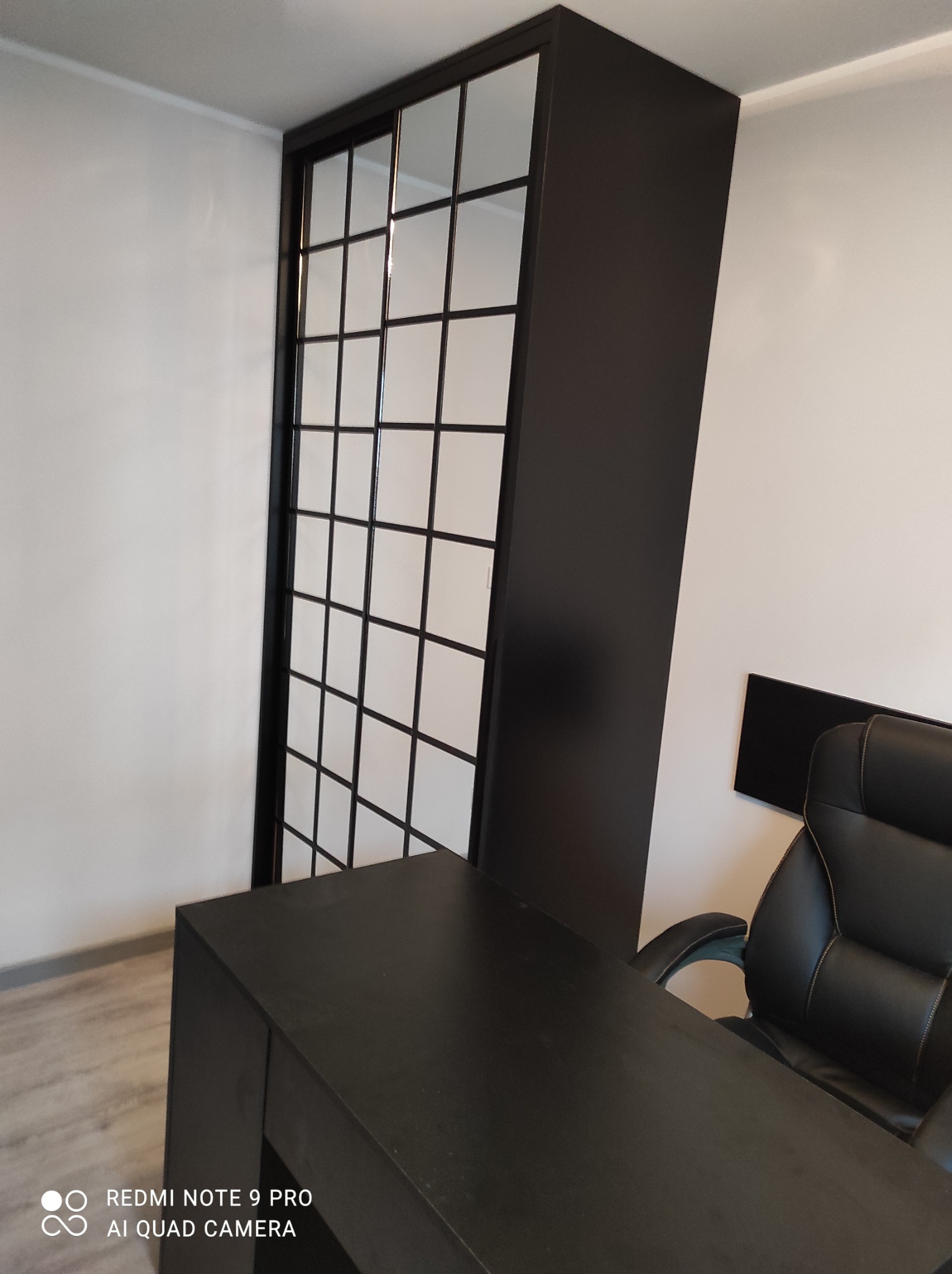 Czarna szafa wnękowa z przesuwnymi drzwiami w kratkę, biurko i fotel w nowoczesnym biurze. Minimalistyczny design, jasne ściany, ciemne meble.