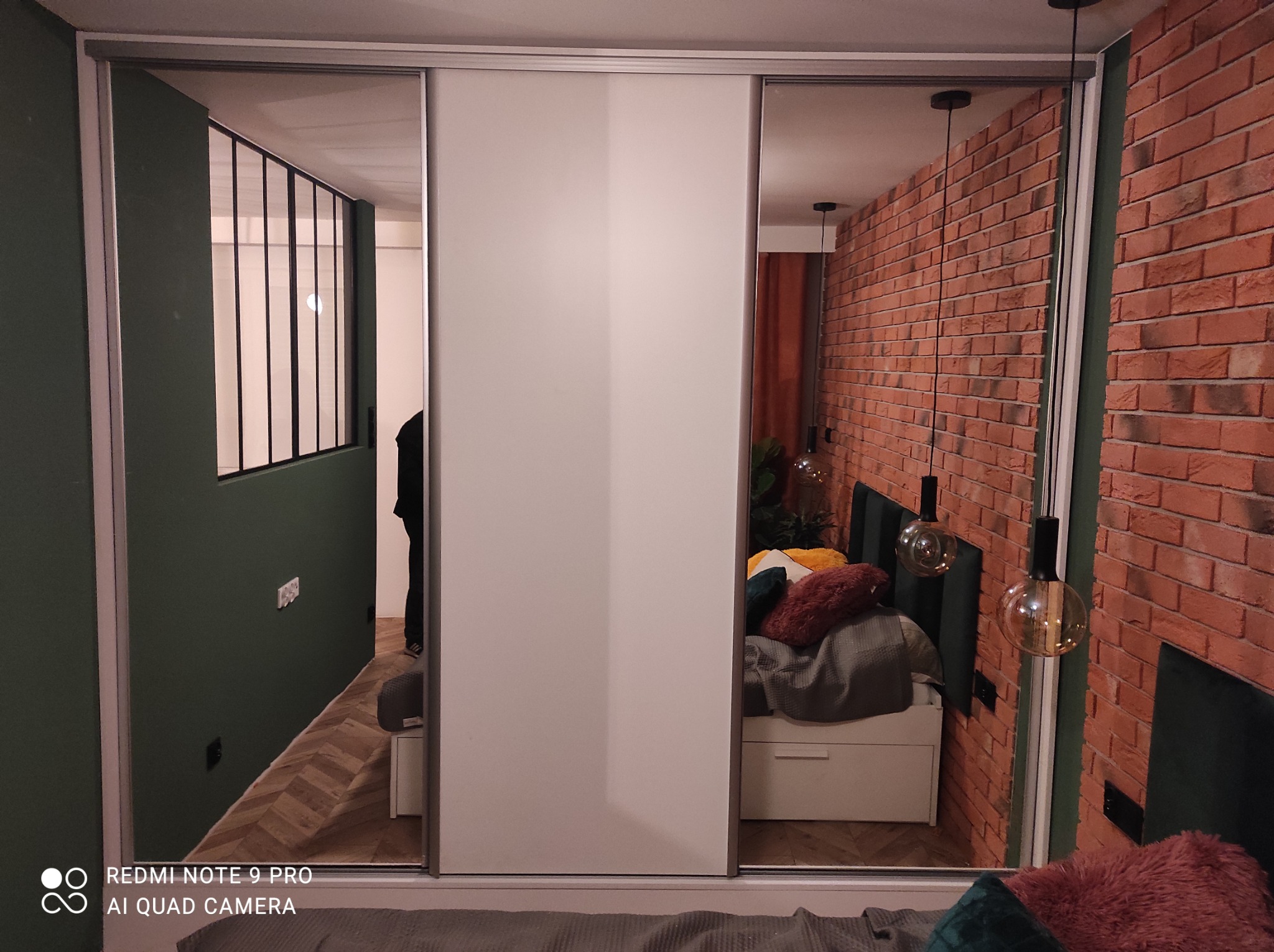 Biała szafa przesuwna z lustrzanymi drzwiami, odbijająca ceglaną ścianę i zieloną ścianę z czarnymi ramami okiennymi w sypialni. Nowoczesny design.