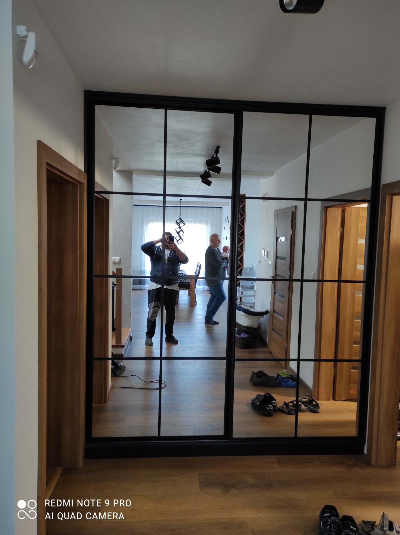 Nowoczesna szafa wnękowa z czarnymi profilami i lustrzanymi drzwiami przesuwnymi, odbijająca wnętrze mieszkania i osoby.