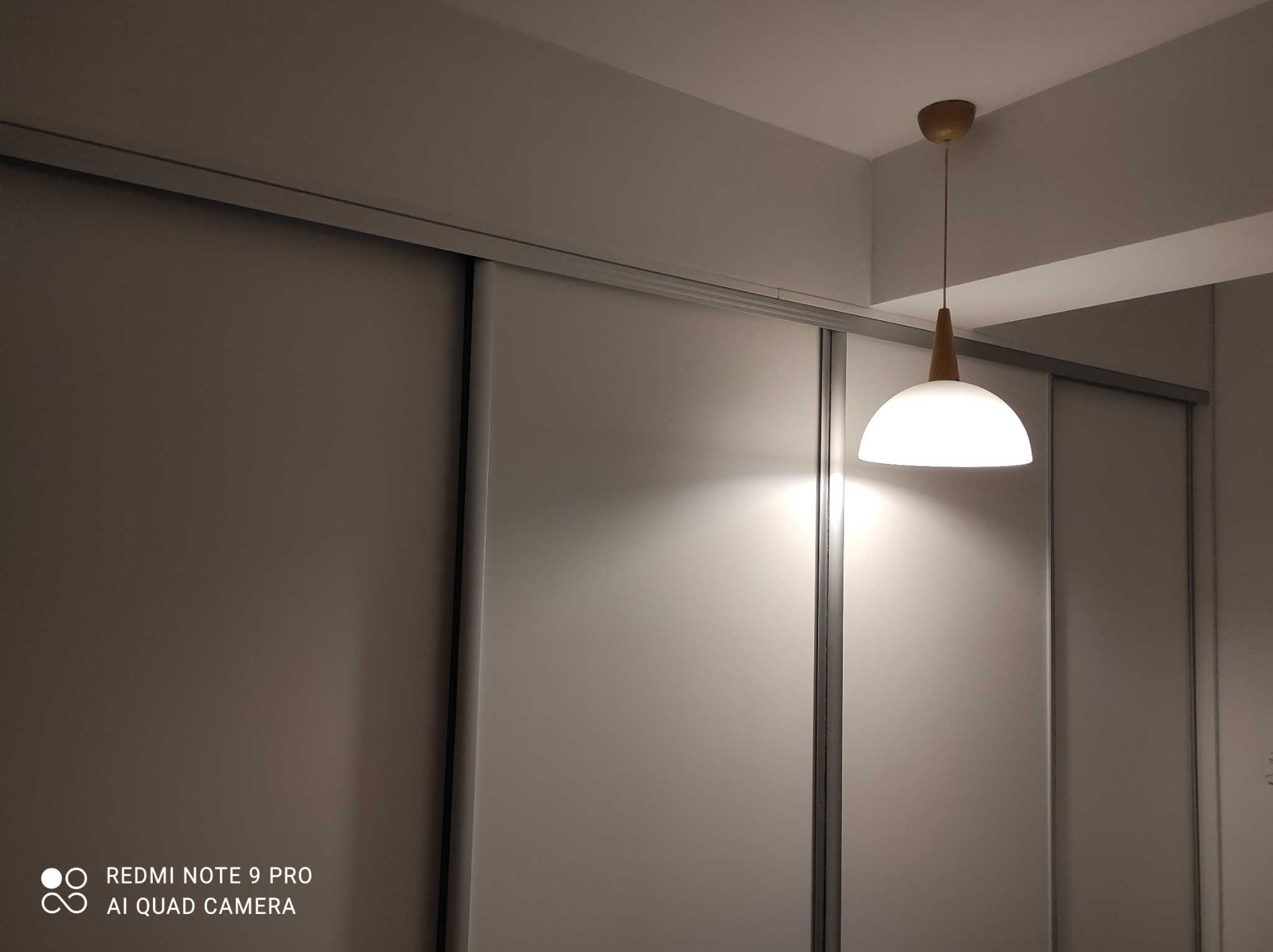 Biała szafa wnękowa z przesuwnymi drzwiami podświetlona lampą wiszącą z drewnianymi elementami. Minimalistyczny design, nowoczesne wykończenie.