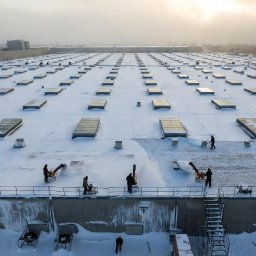 Profesjonalne Odśnieżanie Warszawa - Odśnieżanie dachu hali z świetlikami przez ekipę z użyciem odśnieżarek i łopat. Widok z góry na dużą powierzchnię pokrytą śniegiem i pracujących ludzi.