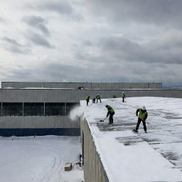 Profesjonalne Odśnieżanie Warszawa - Ekipa w odblaskowych kamizelkach odśnieża płaski dach budynku przemysłowego. Śnieg usuwany łopatami i dmuchawą. Widoczne ślady po odśnieżaniu.