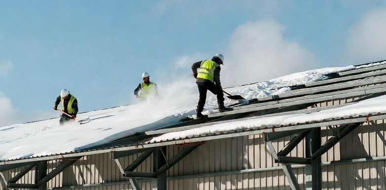 Pracownicy w kaskach i kamizelkach odblaskowych odśnieżają metalowy dach budynku przemysłowego za pomocą łopat. Widoczne chmury pary unoszące się w powietrzu.