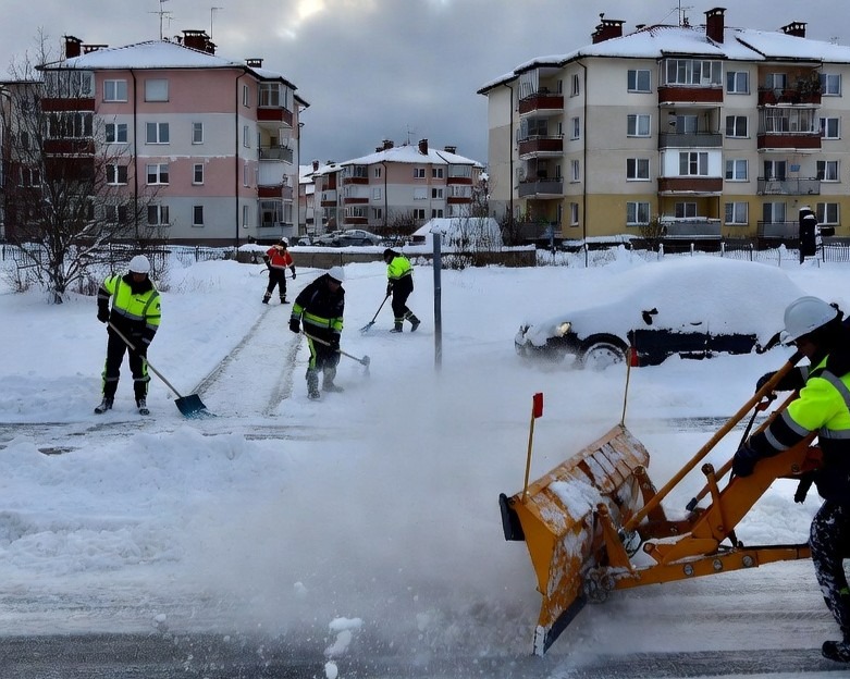Odśnieżanie placu przed blokami przez grupę pracowników w odblaskowych ubraniach, używających łopat i pługu. Samochód zasypany śniegiem w tle.