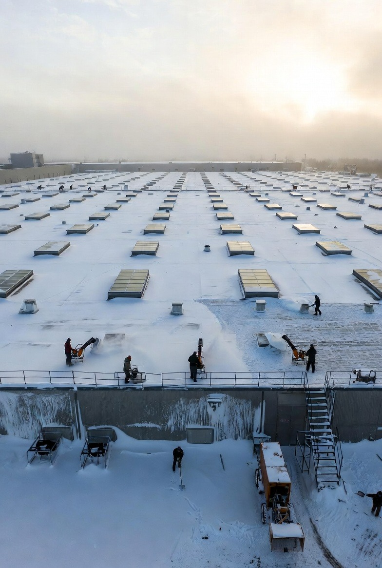 Odśnieżanie dachu hali z świetlikami przez ekipę z użyciem odśnieżarek i łopat. Widok z góry na dużą powierzchnię pokrytą śniegiem i pracujących ludzi.