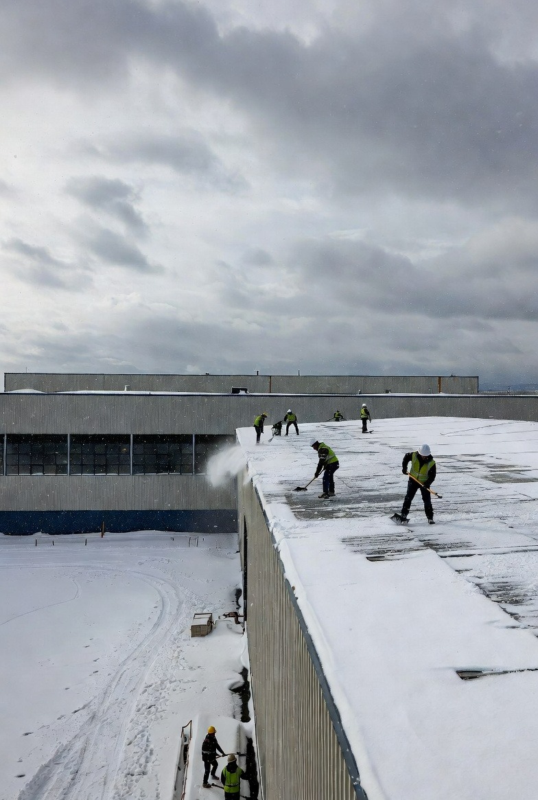 Ekipa w odblaskowych kamizelkach odśnieża płaski dach budynku przemysłowego. Śnieg usuwany łopatami i dmuchawą. Widoczne ślady po odśnieżaniu.