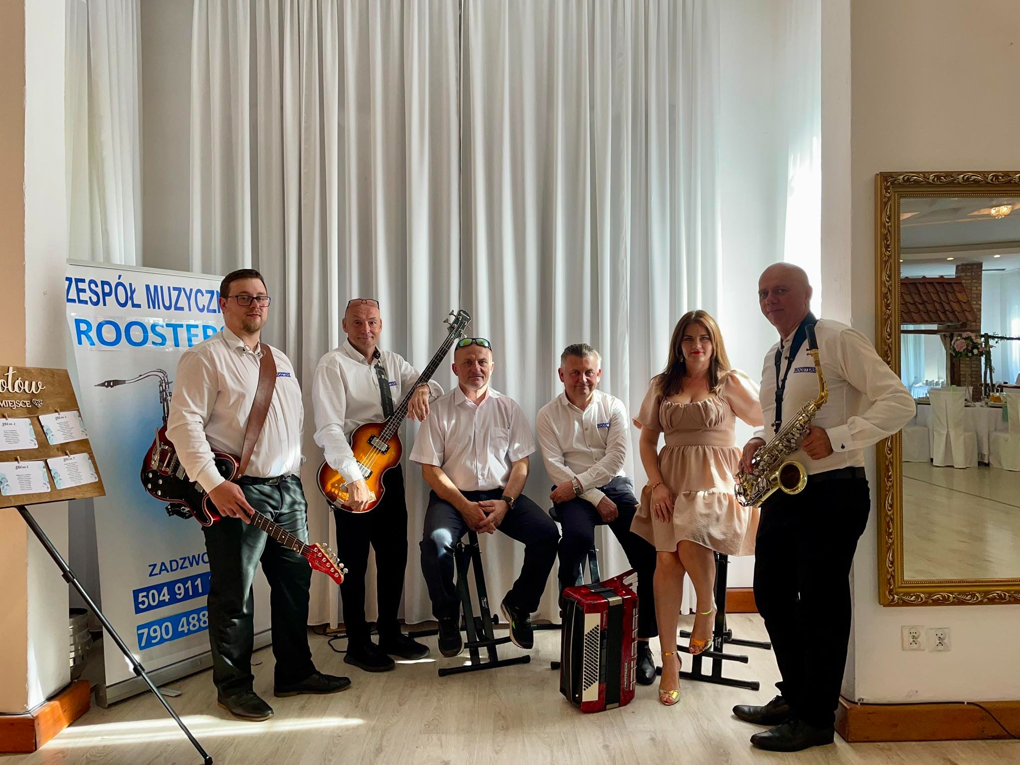 Zespół muzyczny w białych koszulach i z instrumentami (gitary, saksofon, akordeon) pozuje na tle białych zasłon i lustra w złotej ramie. Widoczny plan miejsc.