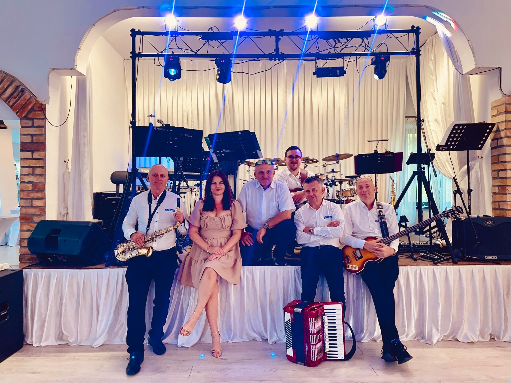 Zespół muzyczny w kompletnym składzie na scenie z instrumentami: saksofon, akordeon, gitara basowa i perkusja, oświetlony niebieskimi reflektorami.