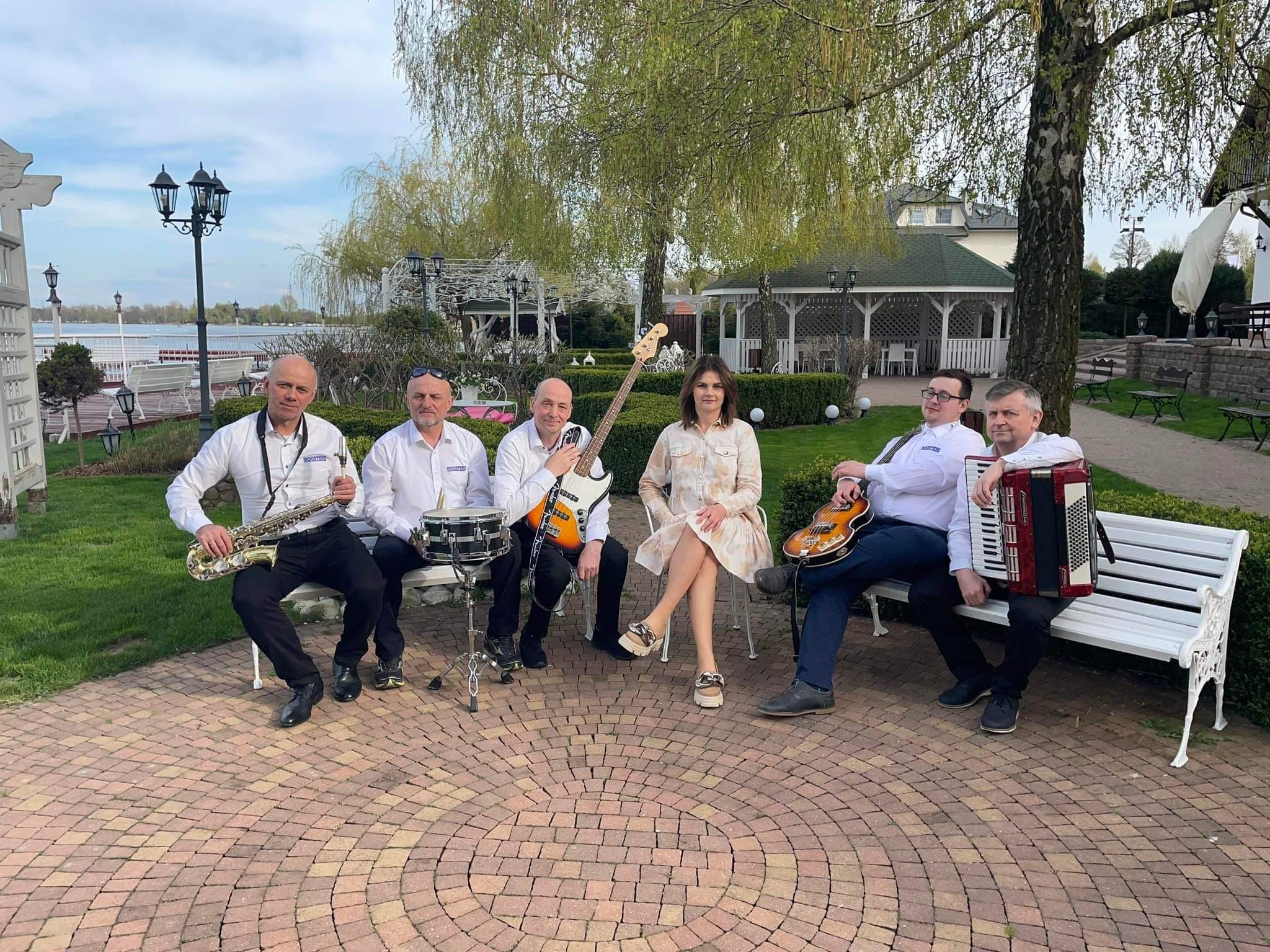 Zespół muzyczny w białych koszulach siedzi na ławce w plenerze z instrumentami: saksofon, gitara basowa, perkusja, gitara elektryczna i akordeon.