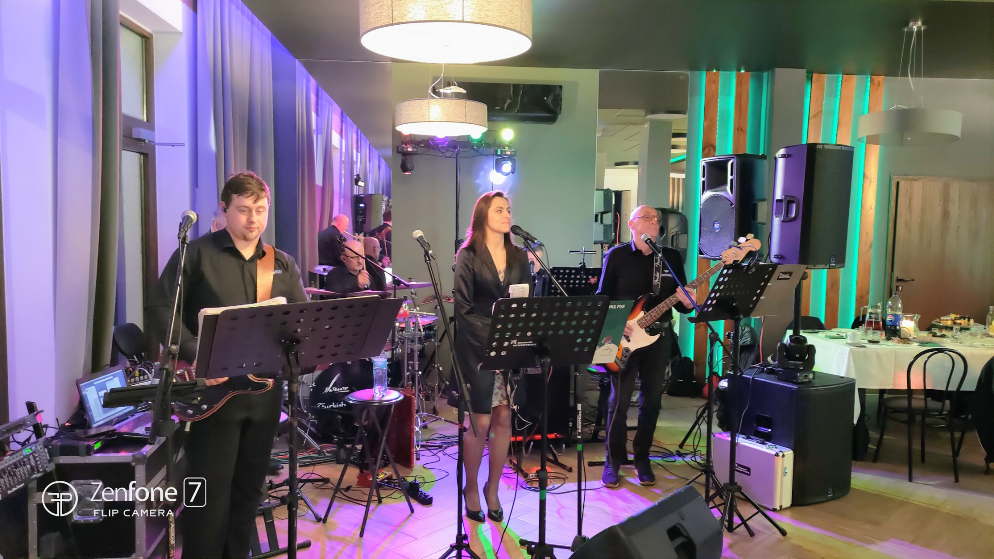 Zespół muzyczny na scenie podczas występu w restauracji. Wokalistka w czarnej marynarce, gitarzyści i perkusista. Oświetlenie sceniczne i nagłośnienie.