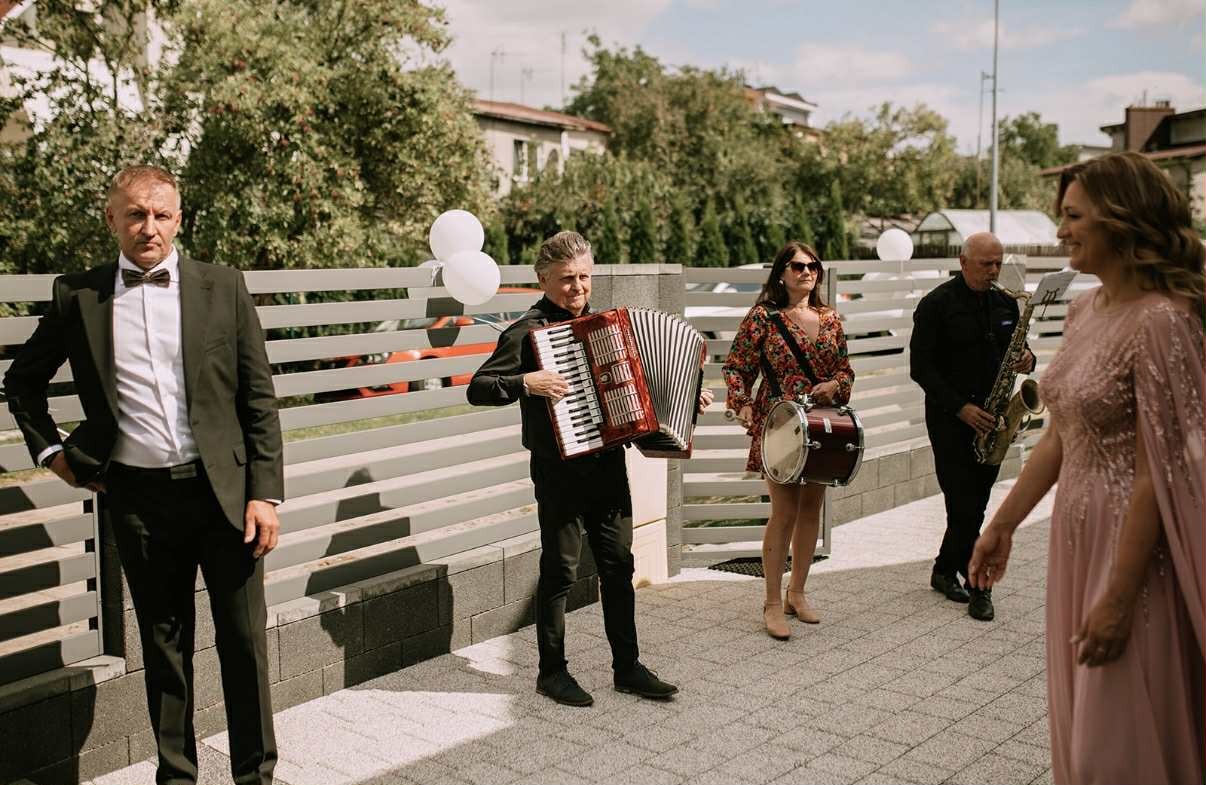 Zespół muzyczny na zewnątrz: akordeonista, perkusistka z bębnem, saksofonista i mężczyzna w garniturze. W tle białe balony i budynki.