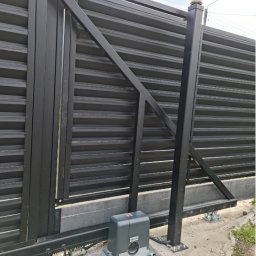 Electric House Tomasz Galoch - Czarna, przesuwna brama z automatyką, widoczny napęd z szarą obudową i żółta lampa ostrzegawcza na słupku. Brama z poziomych profili, zamontowana na betonowej podmurówce.