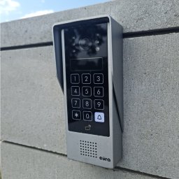 Electric House Tomasz Galoch - Nowoczesny, srebrny wideodomofon z klawiaturą numeryczną i kamerą, zamontowany na szarym, betonowym murze w słoneczny dzień. Widoczny napis 'eura'.