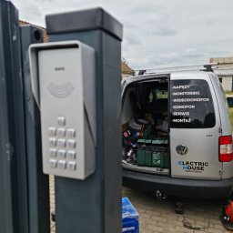 Electric House Tomasz Galoch - Szary domofon z klawiaturą numeryczną zamontowany na metalowym słupku bramy wjazdowej. W tle samochód serwisowy z otwartym bagażnikiem i narzędziami.