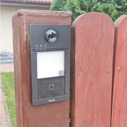 Electric House Tomasz Galoch - Zamontowany wideodomofon HiLook na drewnianym słupku ogrodzenia, z widoczną kamerą, panelem dotykowym i czytnikiem kart zbliżeniowych. Widok z bliska.