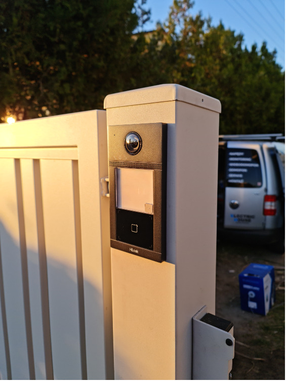 Nowoczesny wideofon HiLook zamontowany na słupku bramy wjazdowej, oświetlony ciepłym światłem zachodzącego słońca. W tle zieleń i samochód dostawczy.
