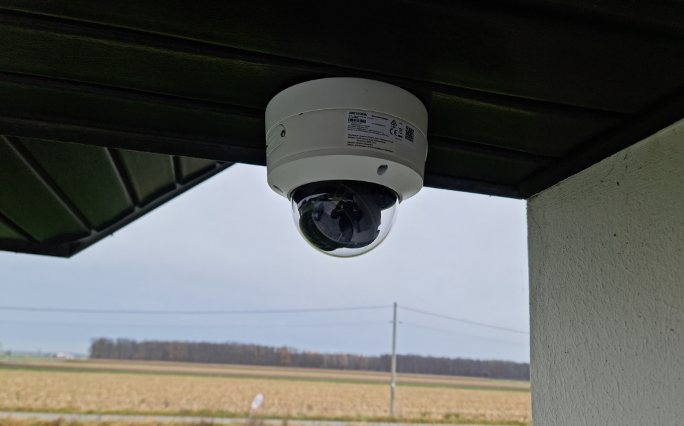 Kamera kopułkowa Hikvision zamontowana pod zadaszeniem, widok z dołu. W tle pole i linia energetyczna. Widoczna etykieta z kodem QR na obudowie kamery.
