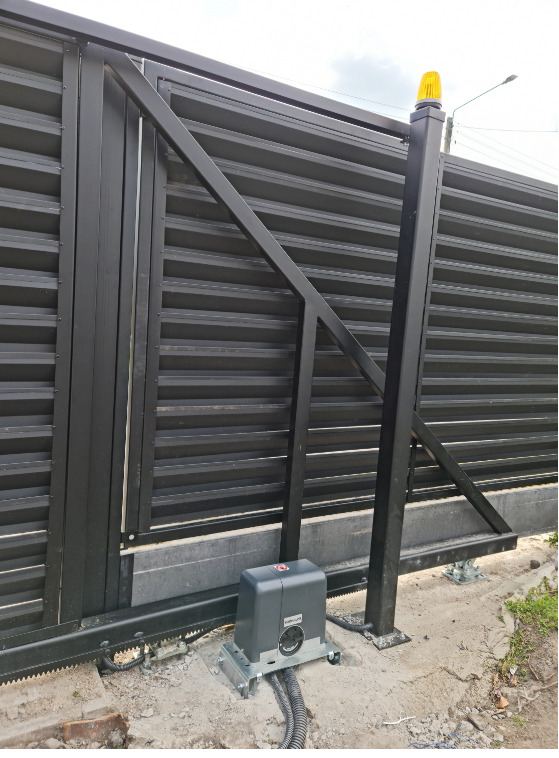 Czarna, przesuwna brama z automatyką, widoczny napęd z szarą obudową i żółta lampa ostrzegawcza na słupku. Brama z poziomych profili, zamontowana na betonowej podmurówce.