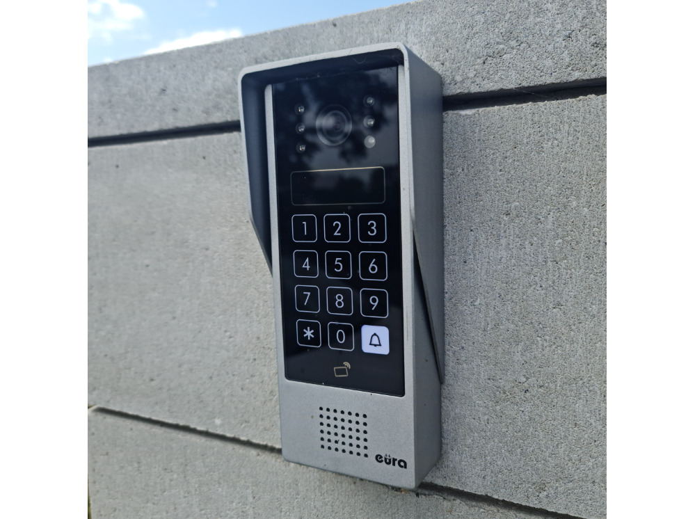 Nowoczesny, srebrny wideodomofon z klawiaturą numeryczną i kamerą, zamontowany na szarym, betonowym murze w słoneczny dzień. Widoczny napis 'eura'.