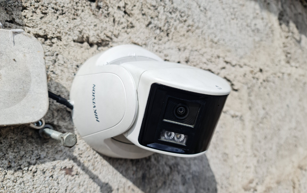 Kamera monitoringu Hikvision, zamontowana na szorstkiej, jasnej ścianie. Widoczny kabel i puszka instalacyjna. Ujęcie z bliska, podkreślające detale urządzenia.