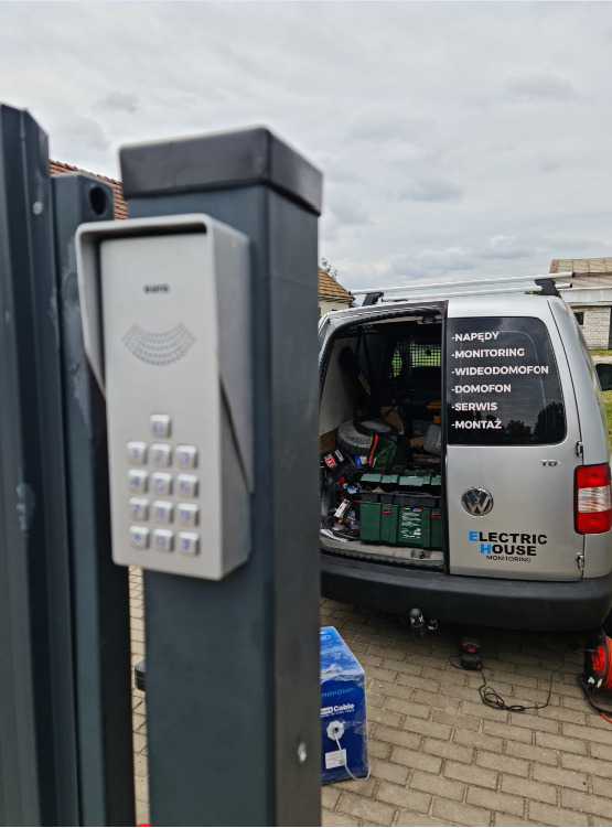 Szary domofon z klawiaturą numeryczną zamontowany na metalowym słupku bramy wjazdowej. W tle samochód serwisowy z otwartym bagażnikiem i narzędziami.