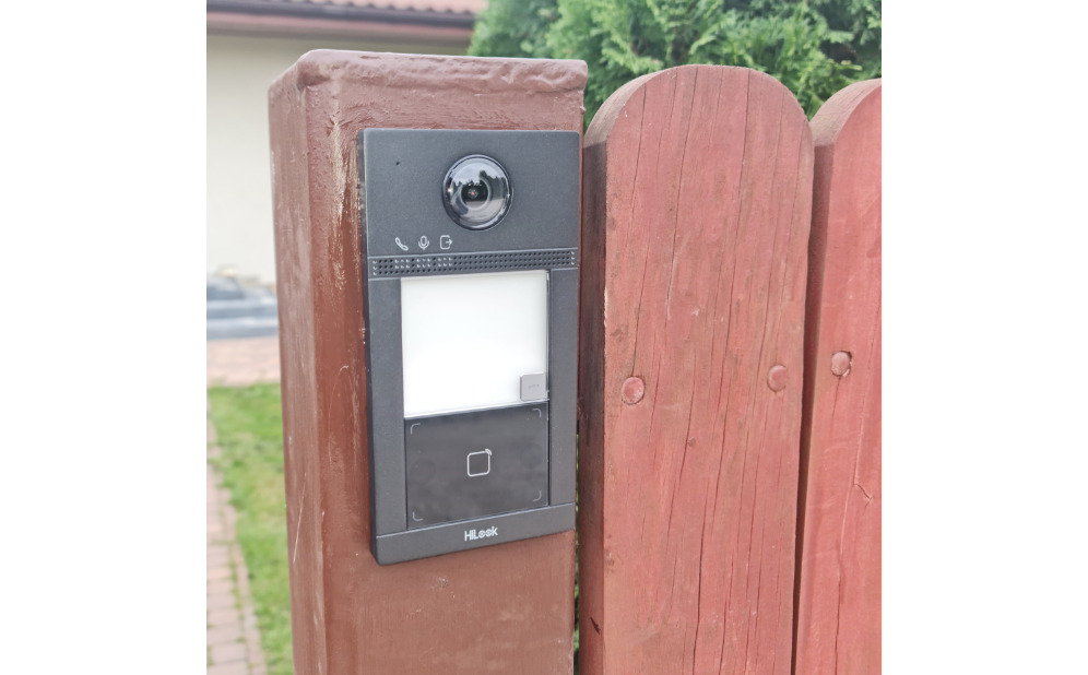 Zamontowany wideodomofon HiLook na drewnianym słupku ogrodzenia, z widoczną kamerą, panelem dotykowym i czytnikiem kart zbliżeniowych. Widok z bliska.