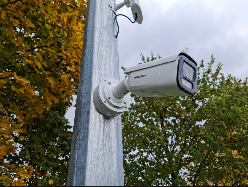 Kamera monitoringu Hikvision zamontowana na metalowym słupie, na tle jesiennych drzew i pochmurnego nieba. Widoczne okablowanie i elementy montażowe.