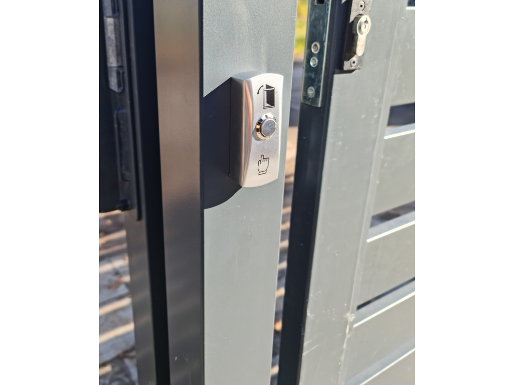 Metalowy panel domofonu z przyciskiem i symbolem dłoni, zamontowany na szarym słupku bramy wjazdowej. Widoczny zamek i fragmenty ogrodzenia.