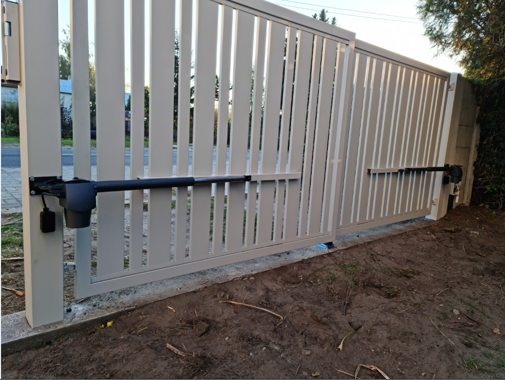 Nowoczesna, przesuwna brama wjazdowa z automatyką, widoczna z bliska. Jasne, pionowe panele bramy i czarne siłowniki. Świeżo wykopana ziemia u podstawy.