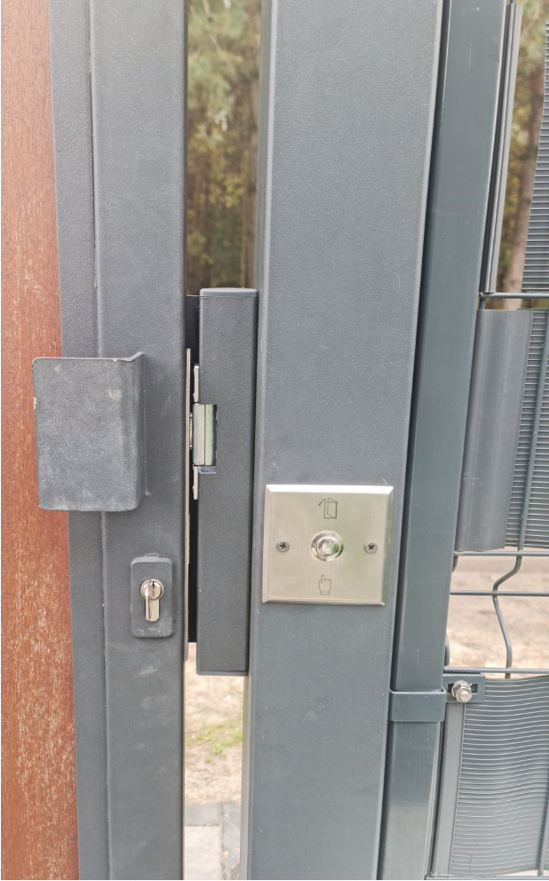 Szczegółowe ujęcie szarej bramy wjazdowej z zamkiem i przyciskiem otwierania, zamontowanego na metalowym słupku. Widoczne elementy konstrukcyjne i otoczenie.