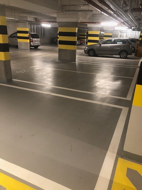 Wnętrze parkingu podziemnego z szarym, lśniącym podłożem, wyznaczonymi białymi liniami miejsc parkingowych i kolumnami w czarno-żółte pasy. Zaparkowane samochody w tle.