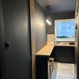 USŁUGI REMONTOWO-BUDOWLANE JAKÓBEK Magdalena Jakóbek - Ciemna kuchnia z drewnianym blatem i hokerami, widok zza ozdobnego portalu z drewnianych listewek. Minimalistyczny design, szare płytki podłogowe.