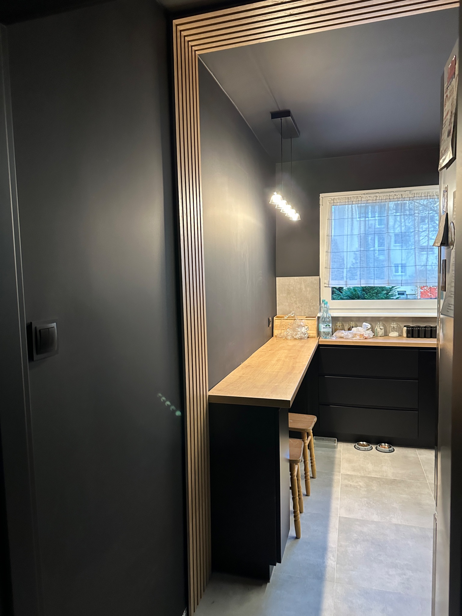 Ciemna kuchnia z drewnianym blatem i hokerami, widok zza ozdobnego portalu z drewnianych listewek. Minimalistyczny design, szare płytki podłogowe.