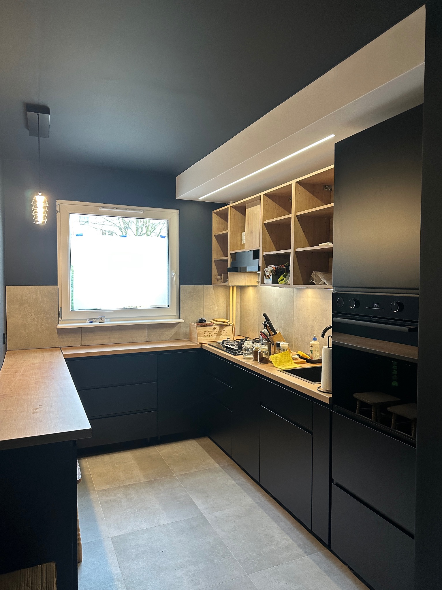 Nowoczesna, czarna kuchnia z drewnianymi blatami i otwartymi półkami, oświetlona listwą LED. Szare płytki podłogowe i ściany w odcieniach grafitu.