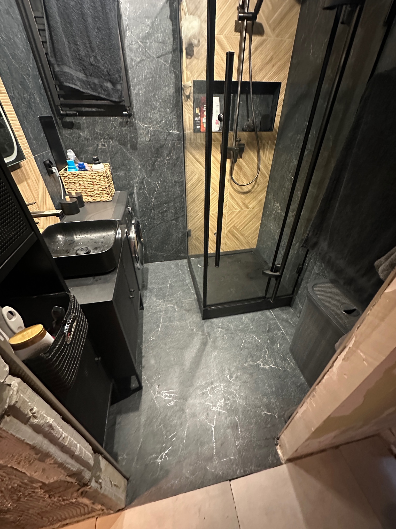 Nowoczesna łazienka z kabiną prysznicową typu walk-in, czarną armaturą i szarą marmurową okładziną. Uwagę zwraca koszyk z akcesoriami obok umywalki.