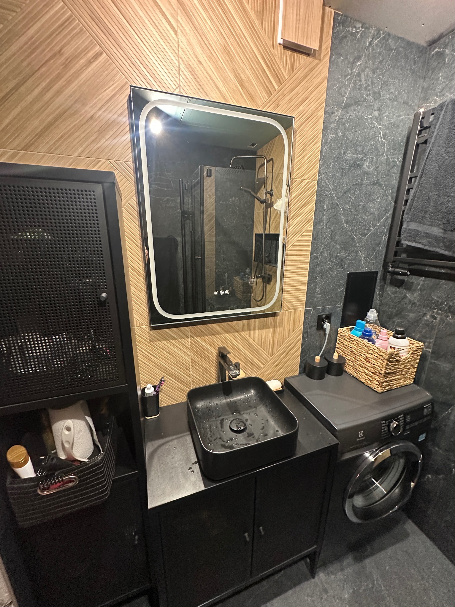 Nowoczesna łazienka z czarną umywalką nablatową, lustrem z podświetleniem LED i pralką. Ściany wyłożone płytkami w odcieniach brązu i szarości. Minimalistyczny design.