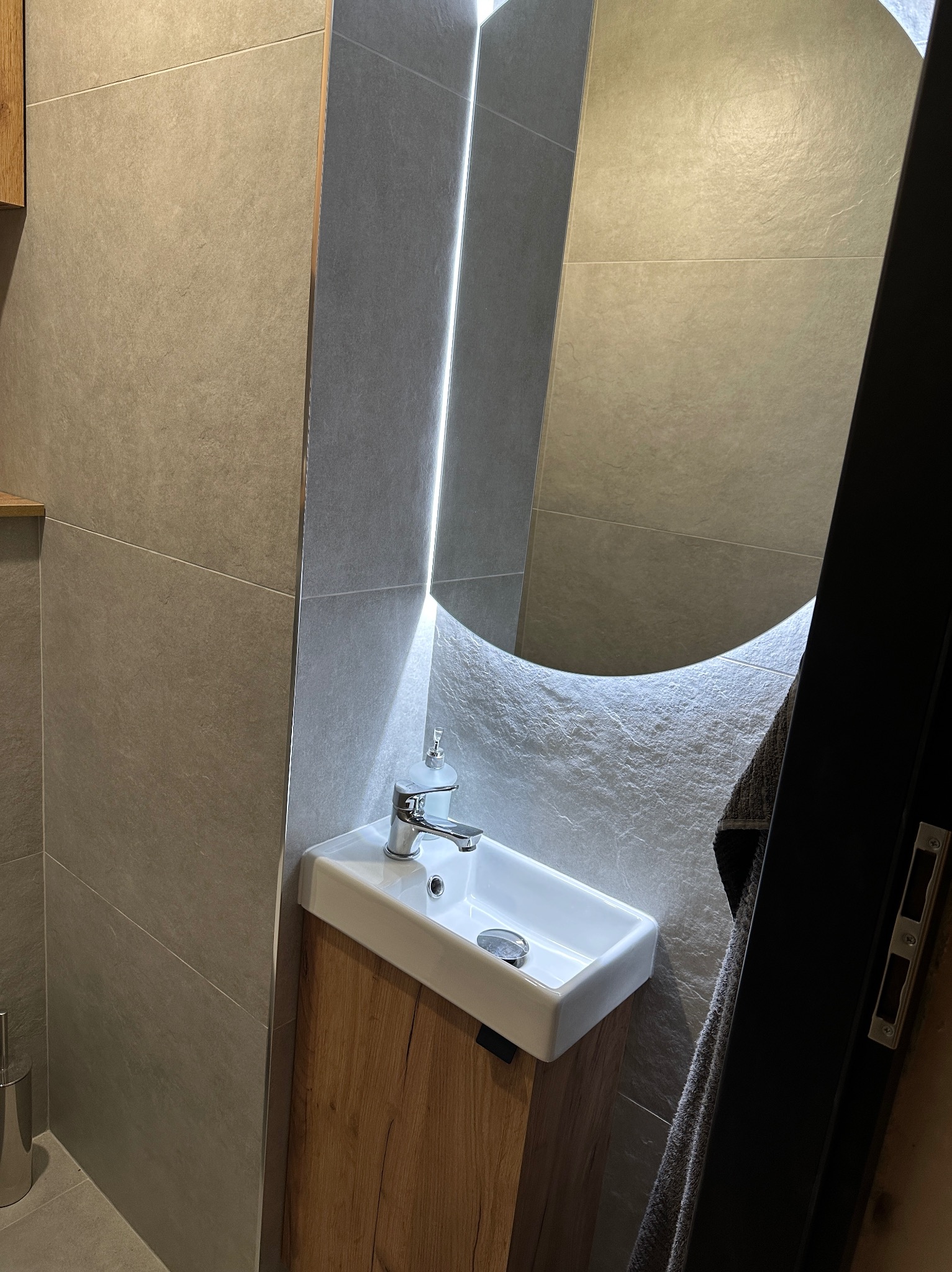 Nowoczesna łazienka z umywalką nablatową na drewnianej szafce, szare płytki na ścianach i okrągłe lustro z podświetleniem LED. Minimalistyczny design.